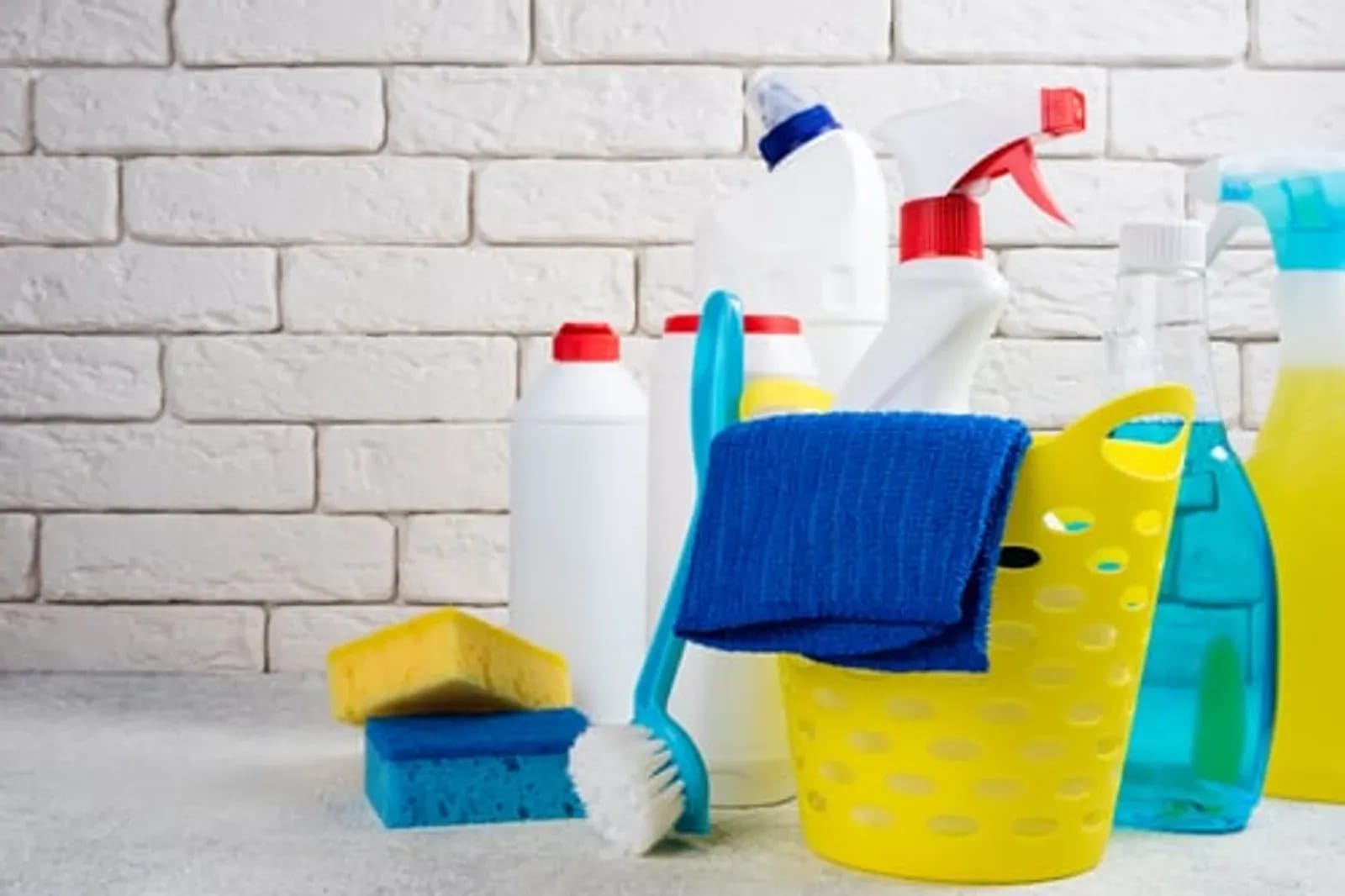 Assorted cleaning bottles, sponges, and a scrub brush arranged against a white brick wall