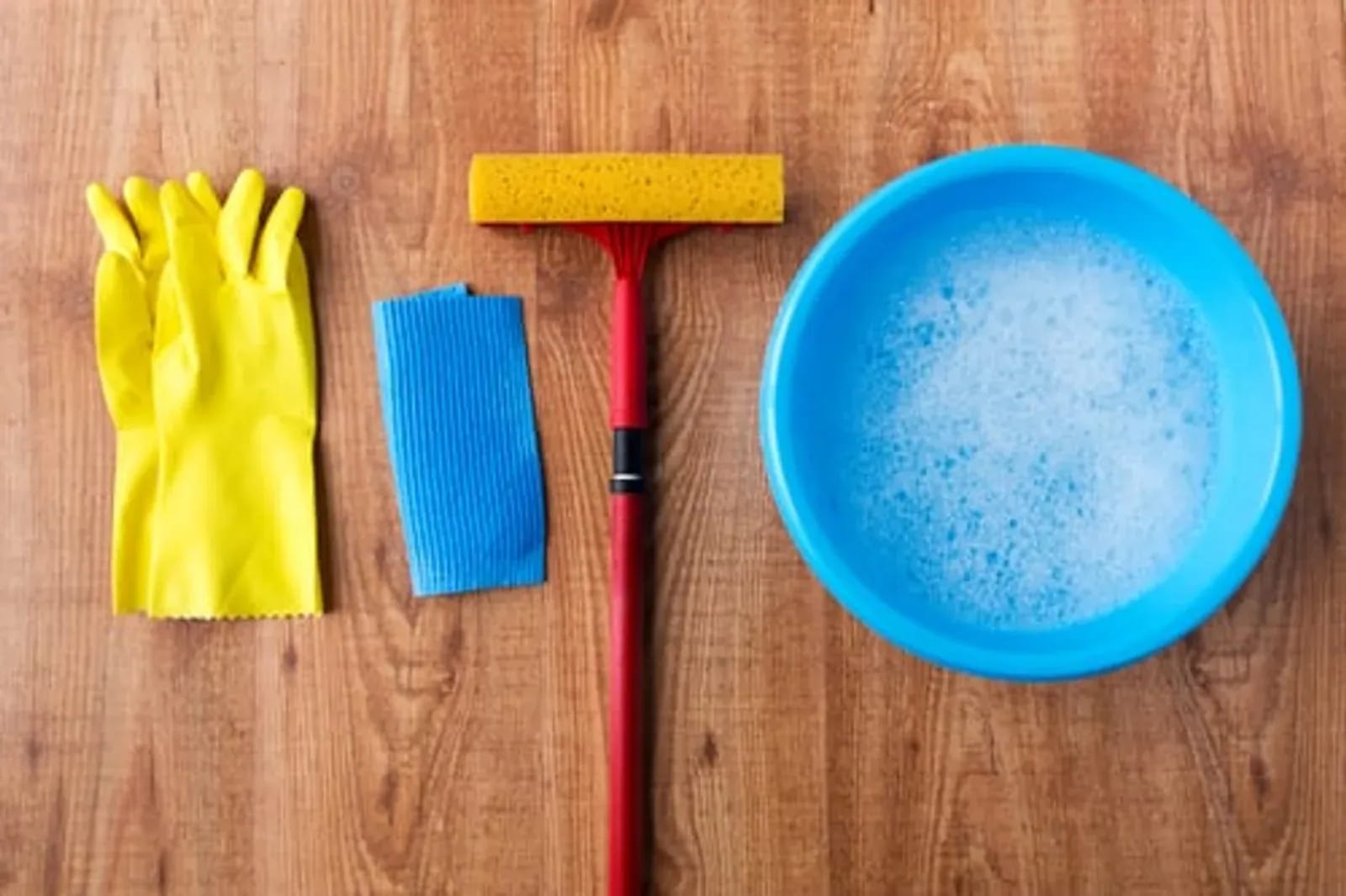Cleaning gloves, cloth, squeegee, and a bowl of soapy water arranged on a wooden floor